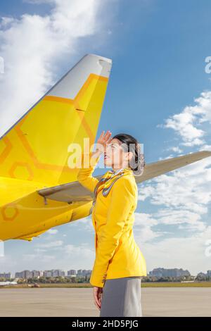 Fröhliche Flugbegleiterin, die mit der Hand die Augen vor der Sonne verhüllte und lächelte, während sie in der Nähe des gelben Flugzeugs am Flughafen stand Stockfoto