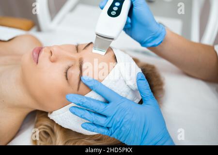 Nahaufnahme einer entspannten Frau erhält eine Ultraschallreinigung der Gesichtsbehandlung durch die Hände eines professionellen Kosmetiks. Peeling der Gesichtshaut in einem Schönheitssalon Stockfoto