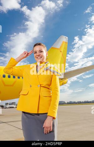 Fröhlicher Flugbegleiter im Anzug der Fluggesellschaft, der in der Nähe eines gelben Flugzeugs steht und mit einer Geste und einem Lächeln lächelt Stockfoto