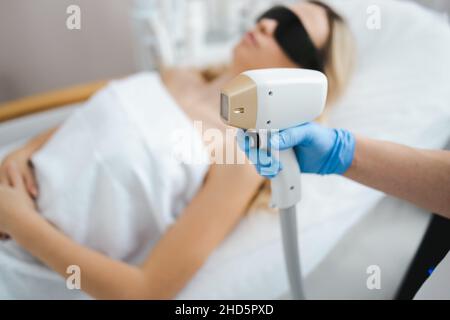 Laserepilierer in der Hand einer Kosmetikerin vor dem Hintergrund einer Patientin, die auf einer Couch liegt. Laser-Haarentfernungskonzept Stockfoto