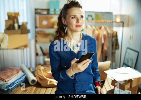 Nachdenkliche elegante Inhaberin eines Kleinunternehmens mittleren Alters mit Smartphone im Büro. Stockfoto