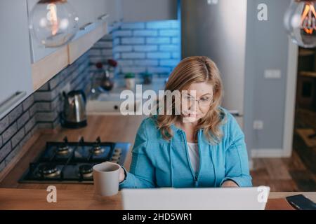 Geschäftsfrau mit blondem Haar trinkt heißen Tee, während sie mit dem Laptop verbunden ist. 40s. Unternehmenstechnologie. Draufsicht. Coronavirus-Zeit. Stockfoto