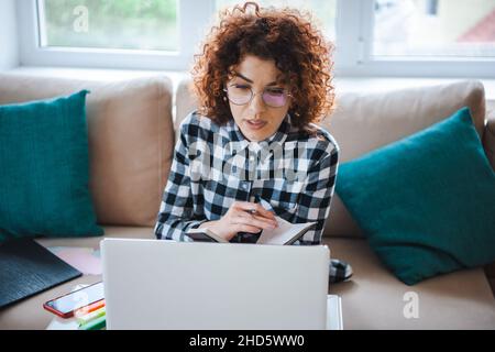 Vorderansicht einer lockigen Frau in einer Brille, die auf dem Sofa sitzt und auf den Laptop-Bildschirm blickt. Fernstudium von zu Hause aus. Freie Mitarbeiterin, die online arbeitet Stockfoto