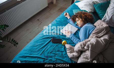 Die obere Ansicht einer lockigen Frau schlief im Bett ein, während sie Hausaufgaben machte, mit einem Notizbuch in der Nähe und einem Stift in der Hand. Stockfoto