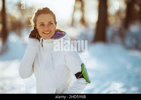 Lächelnde, gesunde Frau in weißer Jacke mit Kopfhörern im Winter im Stadtpark. Stockfoto