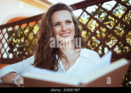 Lächelnde elegante Frau in weißem Hemd mit Buch auf der Terrasse. Stockfoto