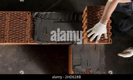 Ein Mann baut eine Backsteinmauer, Bau eines Backsteinhauses. Draufsicht auf den Industriearbeiter, der Keramikblock auf die Zementschicht legt. Stockfoto