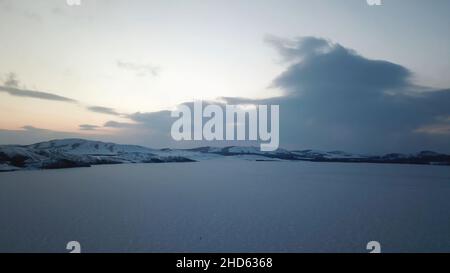 Luftaufnahme Winter Panoramablick auf gefrorenen See umgeben von Hügeln von Bäumen und Schnee bei Sonnenuntergang wolkigen Himmel Hintergrund. Fliegen über eisigen See oder Pon Stockfoto