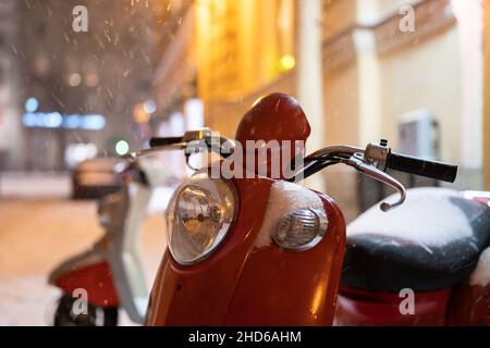 Roter Retro Vintage Motorroller unter fallendem Schnee, der nachts auf der winterlichen Stadtstraße geparkt ist Stockfoto