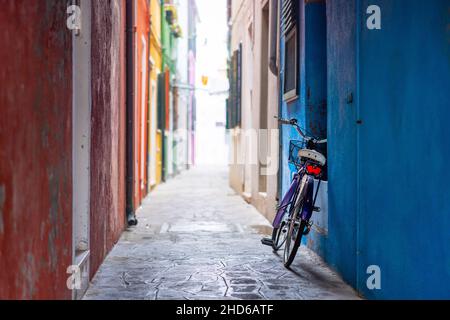 In einer bunten Hintergasse in Burano wird ein violettes Fahrrad zurückgelassen, das sich an einer blauen Wand lehnt Stockfoto