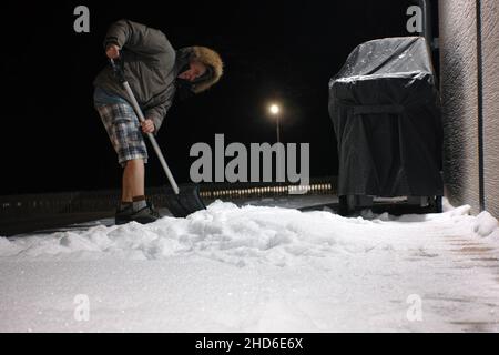 Ein Mann mit Schaufel, der Schnee um ein Haus entfernt, Nachtszene, Fokus im Vordergrund Stockfoto