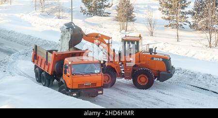 23. Dezember 2021. Region Kemerowo, Russland. Großer orangefarbener Traktor reinigt Schnee von der Straße und lädt ihn in den LKW. Reinigung und Reinigung der Straße Stockfoto