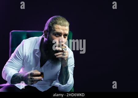 Porträt eines hübschen bärtigen Mannes, der Zigarre raucht, hält ein Glas Whiskey in einem Sessel. Stockfoto