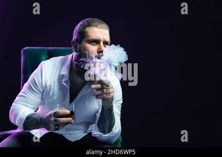 Porträt eines bärtigen Hipster-Mannes, der Zigarre raucht, hält ein Glas Whiskey in einem Sessel. Stockfoto