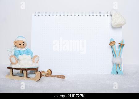 Teddybär in winterblauer Kleidung auf einem Schlitten, leuchtend blaue Holzskier, weißer Hut und leeres Notizbuch auf weißem Schnee. Wintersport-Konzept. Sport Stockfoto