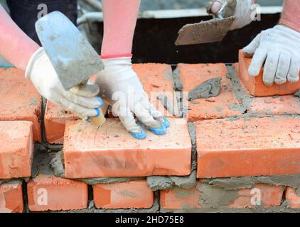 Zwei Maurer legen Ziegel aus einer Ziegelwand, die mit einer Maurerkelle und Mörtel untermauert werden. Nahaufnahme eines Mauerwerks aus verbrannten Ziegelsteinen. Stockfoto