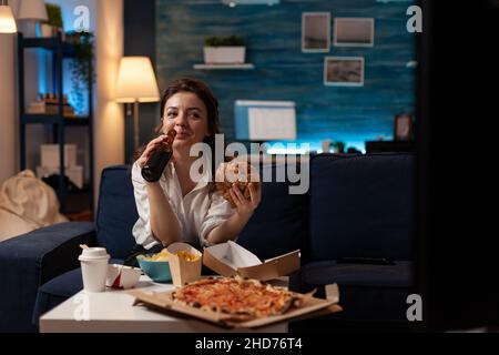Kaukasische Frau trinkt Craft-Flaschenbier und isst einen köstlichen Hamburger zum Mitnehmen, der auf der Couch im Wohnzimmer sitzt. Person, die vor dem Fernseher lacht und Pizza und Junk Food zum Mitnehmen bestellt hat. Stockfoto