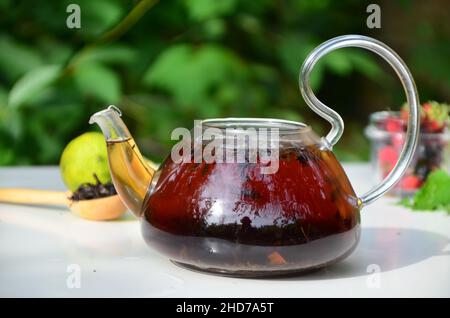 Teekannen voll Tee auf einem Hintergrund von grünem Laub auf einem Holztisch, neben Waldbeeren, Himbeeren und Heidelbeeren, Limette oder Bergamotte, Minze. Er Stockfoto
