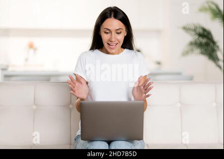 Kaukasische junge Erwachsene schöne glückliche Brünette Frau, die im Wohnzimmer mit einem Laptop auf dem Sofa sitzt, über eine Videokonferenz spricht, mit den Händen gestikelt, auf den Laptop-Bildschirm blickt und freundlich lächelt Stockfoto