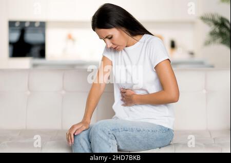 Frustrierte junge kaukasische Frau, die auf dem Sofa im Wohnzimmer sitzt, Bauchschmerzen, Gesundheitsprobleme, Krämpfe, Geschwüre, Probleme mit dem Verdauungstrakt, brauchen eine ärztliche Beratung Stockfoto