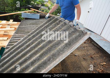 Ausbau und Austausch des Dachs aus altem Asbestzement Ein Dachdecker entfernt alte Asbest-Dachbleche während der Dachsanierung. Dachkonstruktion Stockfoto