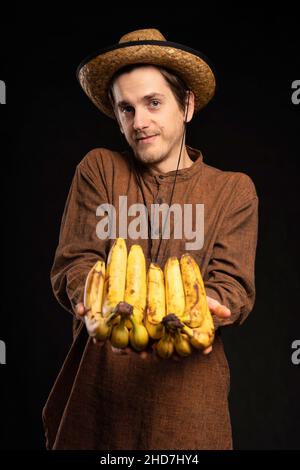 Junger, hübscher, großer, schlanker weißer Mann mit braunen Haaren, der Bananen mit braunem Hemd und Strohhut auf schwarzem Hintergrund anbietet Stockfoto