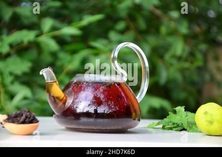 Teekannen voll Tee auf einem Hintergrund von grünem Laub auf einem Holztisch, neben Waldbeeren, Himbeeren und Heidelbeeren, Limette oder Bergamotte, Minze. Er Stockfoto