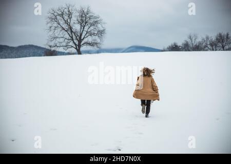 Frau in braunem Mantel läuft auf einem Schneefeld. Berge in der Ferne. Free Spirit Girl. Wintersaison. Hochwertige Fotos Stockfoto