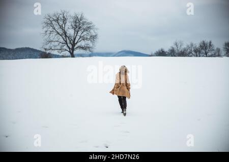 Frau in braunem Mantel läuft auf einem Schneefeld. Berge in der Ferne. Free Spirit Girl. Wintersaison. Hochwertige Fotos Stockfoto