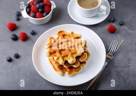 Wiener oder belgische Waffeln mit frischen Beeren (Himbeeren und Blaubeeren) auf einem weißen Teller und einer Tasse Kaffee. Traditionelles Dessert. Nahaufnahme. Stockfoto