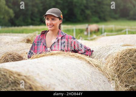 Junge Frau in Hemd und Mütze lächelnd, Heustapel rollt neben ihr Stockfoto