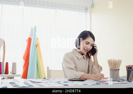 Schöne asiatische Designerin, die am Telefon mit dem Kunden spricht und die Arbeitsdetails zur Kenntnis nimmt, um den Auftrag für den Schneider zu erhalten Stockfoto