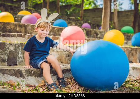 Niedlicher kleiner Junge mit Hasenohren, der sich mit der traditionellen Ostereiersuche im Freien amüsieren kann. Wir feiern die Osterferien. Kleinkinder finden, bunte Eier Stockfoto