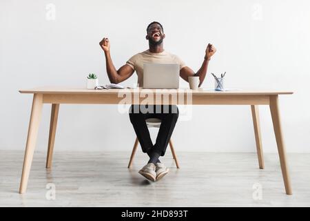 Aufgeregt junger schwarzer Mann, der VOR dem Laptop JA gestikuliert, Erfolg oder Erfolg gefeiert und im Büro am Schreibtisch sitzt Stockfoto