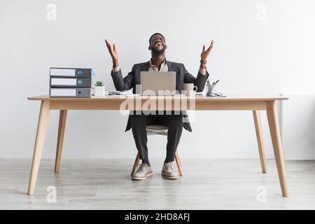In voller Länge Porträt eines überglücklich jungen afroamerikanischen Geschäftsmannes, der mit einem Laptop an einer weißen Wand am Tisch sitzt Stockfoto