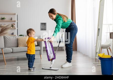 Eine glückliche Familie, die das Zimmer sauber gemacht hat. Mutter und Kleinkind Sohn tun die Reinigung zu Hause, Kind Junge hilft Mutter waschen Boden Stockfoto