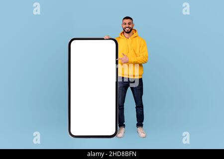 Glücklicher arabischer Mann, der auf ein großes Smartphone mit weißem, leeren Bildschirm zeigt und freien Kopierplatz für Ihr Design zeigt Stockfoto