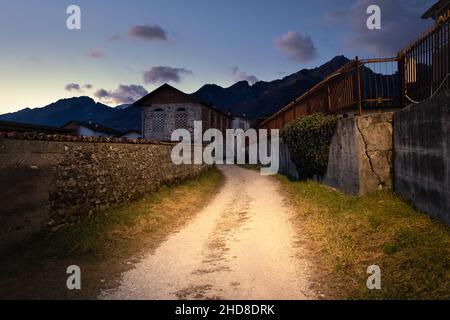 Feldweg zwischen alten Häusern bei Sonnenuntergang, Berge im Hintergrund, Santa Giustina, Belluno Stockfoto