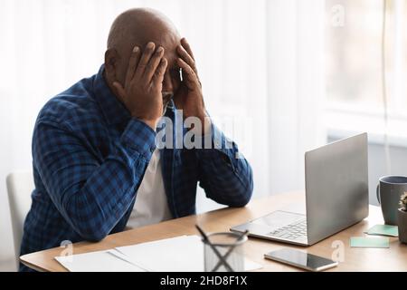 Unglücklicher Älterer Schwarzer Geschäftsmann, Der Das Gesicht Bedeckt, Das In Der Nähe Des Laptops In Der Halle Sitzt Stockfoto