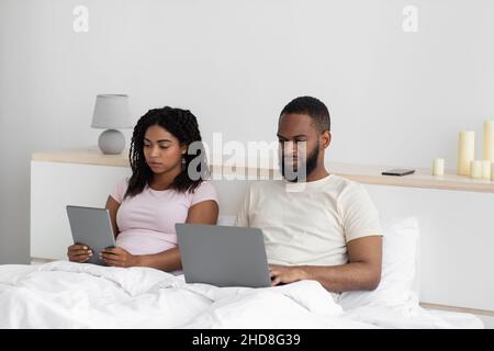 Beschäftigt ernsthafte Millennial schwarze Frau und Mann arbeiten auf Laptop und Tablet, sitzen auf dem Bett Stockfoto