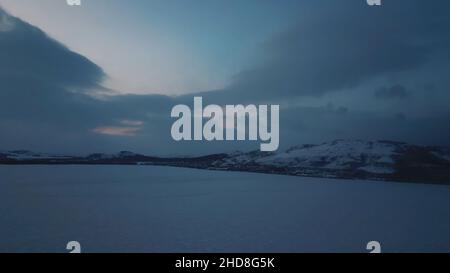 Luftaufnahme Winter Panoramablick auf gefrorenen See umgeben von Hügeln von Bäumen und Schnee bei Sonnenuntergang wolkigen Himmel Hintergrund. Fliegen über eisigen See oder Pon Stockfoto