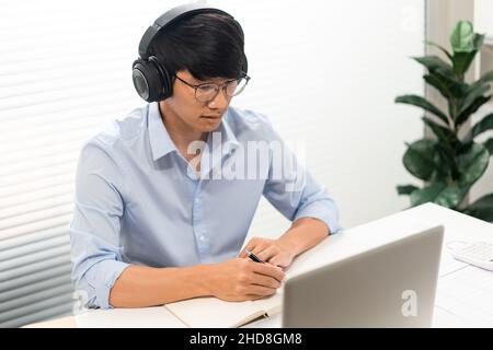 Working man Conept der männliche Offizier, der sich auf den Laptop konzentriert, etwas auf das Papier schreibt, während er Kopfhörer trägt. Stockfoto