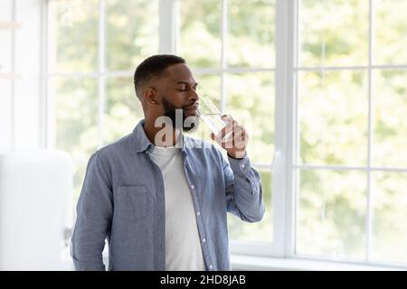 Junger hübscher Schwarzer mit Bart trinkt sauberes Wasser aus Glas mit geschlossenen Augen am Fensterhintergrund Stockfoto