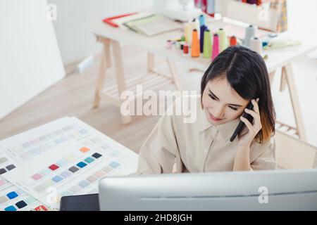 Schöne asiatische Designerin, die am Telefon mit dem Kunden spricht und die Arbeitsdetails zur Kenntnis nimmt, um den Auftrag für den Schneider zu erhalten Stockfoto