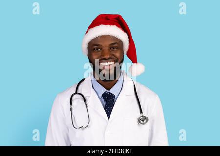Schöner schwarzer Arzt in Uniform und Weihnachtsmann Hut posiert auf blauem Hintergrund Stockfoto