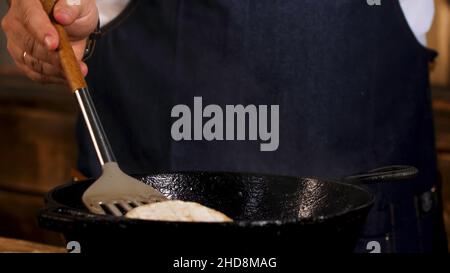 Nahaufnahme eines männlichen Köchers, der Camembert-Käse in einer schwarzen Eisenpfanne von beiden Seiten mit einer Metallschaufel braten soll. Prozess der Zubereitung von Speisen im Restaurant Stockfoto