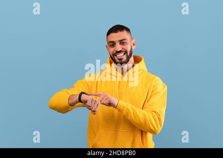 Fröhlicher Mann aus dem Nahen Osten, der seine Smartwatch berührt und die Kamera anlächelt und über dem blauen Hintergrund des Studios steht Stockfoto