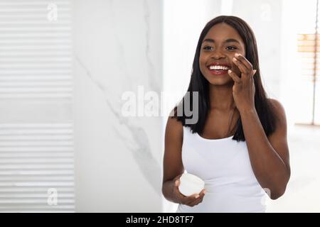 Startseite Beauty-Routine. Schöne Schwarze Frau Anwendung Feuchtigkeitsspendende Creme Auf Gesicht Im Badezimmer Stockfoto