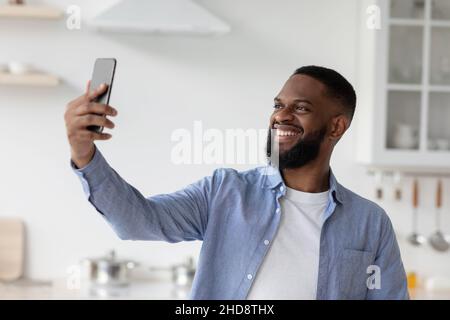 Zufriedener Tausendjähriger afroamerikanischer Mann mit Bart macht Foto am Telefon im Kücheninnenraum Stockfoto