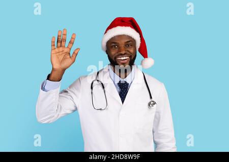 Lächelnder Schwarzer Doktor In Weihnachtsmütze Winkt Hand An Kamera Stockfoto